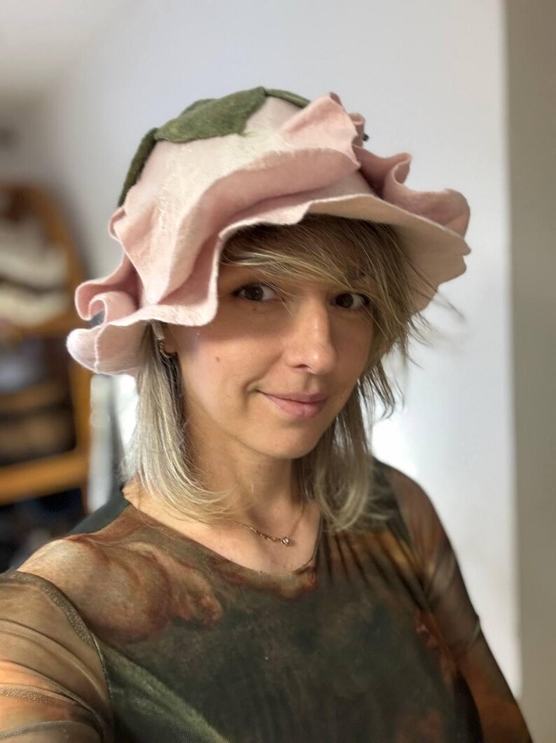Pode incluir: Um chap&eacute;u artesanal &uacute;nico em forma de rosa rosa grande, com folhas verdes e uma aba com babados. Feito de material de feltro macio, este acess&oacute;rio artesanal &eacute; ideal para fantasias ou eventos especiais, adicionando um toque floral.
