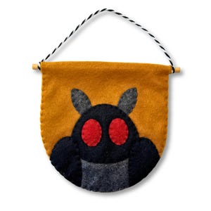 Può includere: Un banner di feltro con un design di creature dei cartoni animati. Lo striscione ha uno sfondo giallo senape con una creatura nera con occhi rossi. La creatura ha orecchie e braccia grigie. Un tassello di legno e uno spago bianco e nero in alto.