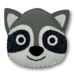 May include: A handmade felt raccoon face with gray, white, and black details. The raccoon has button eyes and a stitched smile. The ears are gray with white inner ears. The edges are stitched with gray thread.