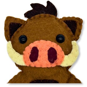 Pode incluir: Um javali de feltro feito à mão com corpo castanho, detalhes castanhos escuros e focinho laranja claro. O javali tem olhos de botão pretos, presas amarelas claras e é costurado com linha castanha. O design é em estilo cartoon.