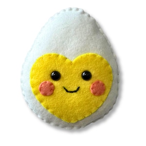 May include: A white felt egg-shaped plush toy with a yellow heart-shaped center. The heart has black button eyes, a stitched smile, and pink blush circles. The egg has a scalloped edge.