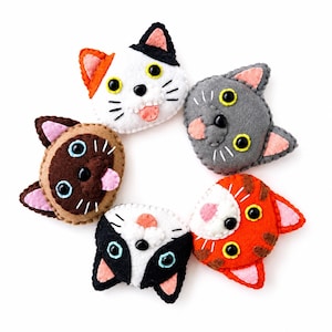 May include: Five felt cat head ornaments arranged in a circle. The cats feature various colors including white with black and orange patches, solid gray, brown, black and white, and orange tabby. Each has stitched details and button eyes.