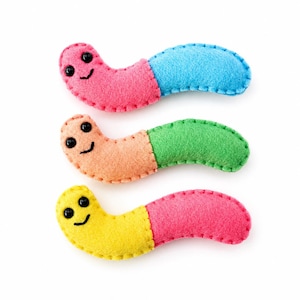 May include: Three felt worm toys, each featuring a two-tone design with stitched edges and black bead eyes. The top worm is pink and blue, the middle is peach and green, and the bottom is yellow and pink. Each has a stitched smiling mouth.