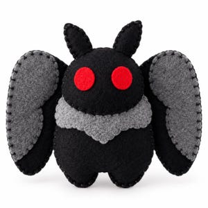 Può includere: Un peluche in feltro fatto a mano a forma di falena, con un corpo nero, due occhi circolari rosso brillante e grandi ali grigie. I bordi dei pezzi in feltro sono uniti tra loro con cuciture nere visibili che creano un contorno.