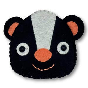 May include: A handmade felt skunk face with a black body, white stripe, and orange accents. The skunk has button eyes, an orange nose, and a stitched smile. The ears are orange inside. This item is likely a decorative piece or a toy.