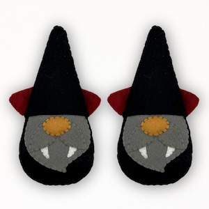May include: Two felt vampire gnome ornaments. Each gnome has a black pointed hat, a grey face with a yellow nose, and two white fangs. The gnomes have red accents on their hats and black bodies.