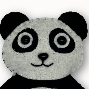 May include: A handmade panda bear face made of gray and black felt. The panda has black button eyes, a black nose, and a stitched smile. The edges are stitched with white thread, and the ears are black. This item is a craft or toy.