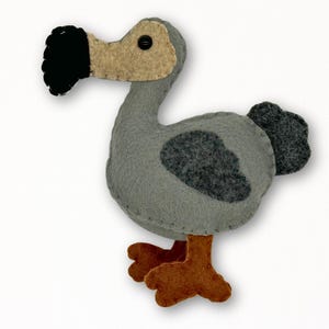 Pode incluir: Um brinquedo de feltro feito à mão de um pássaro dodô em tons de cinza, bege, marrom e preto. O pássaro tem um bico preto, um bico e cabeça bege e um corpo cinza com uma mancha cinza mais escura. Os pés são marrons e o olho é um botão preto.