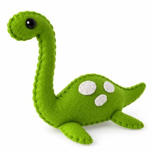 Può includere: Un peluche a forma di dinosauro in feltro verde brillante con un collo lungo e un corpo arrotondato. La creatura presenta un occhio a perla nera, cuciture visibili lungo i bordi e tre piccole toppe circolari in feltro bianco sul fianco.