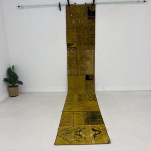 Puede incluir: Una alfombra larga y rectangular de patchwork en tonos verde oliva y marrón. La alfombra presenta una variedad de patrones y texturas geométricas, creando una estética vintage o bohemia. La alfombra se exhibe sobre un suelo blanco.
