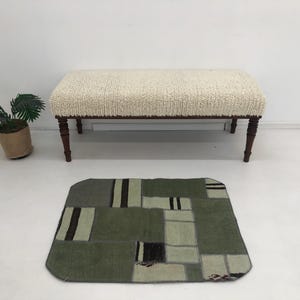 以下が含まれることがあります： 濃い茶色の脚と長方形のテクスチャード加工の座面を備えた、クリーム色のベンチ。緑とグレーのパッチワークラグがベンチの前にあります。鉢植えの植物がベンチの左側にあります。