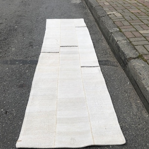 Könnte beinhalten: Ein weißer gewebter Teppich mit schwarzen Nähten. Der Teppich ist rechteckig und hat ein schlichtes Design.