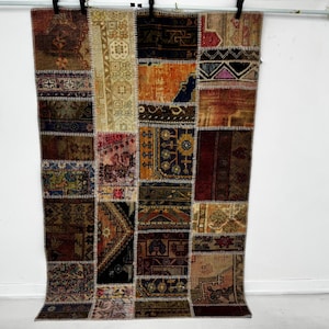 May include: A patchwork rug with a variety of colours and patterns. The rug is made up of rectangular pieces of fabric stitched together. Colours include brown, beige, orange, and burgundy. The rug is hanging against a white wall.