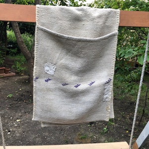 以下が含まれることがあります： 手作りの薄いベージュ色の生地の吊り下げポケット。ステッチされたボーダーがあり、パッチと紫色の刺繍デザインがあります。白いロープが付いた木製のブランコフレームから吊り下げられています。