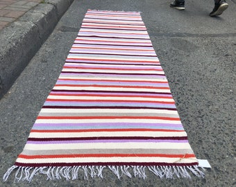 May include: A long, rectangular, striped rug with a white background and alternating stripes of orange, red, purple, light blue, and gray. The rug has white fringe on both ends.
