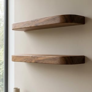 Könnte beinhalten: Zwei schwebende Holzregale mit einer gebogenen Vorderkante sind an einer beigefarbenen Wand montiert. Die Regale sind aus dunkelbraunem Holz gefertigt. Die Regale sind leer. Dekorative Vasen und Bücher befinden sich auf einem unteren Regal.