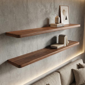 May include: Two wooden floating shelves with a natural wood grain are mounted on a grey concrete wall. The top shelf holds a framed print, a vase with branches, and books. The bottom shelf has books. The shelves are above a beige sofa.