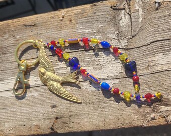 May include: A gold-colored bird charm keychain with a beaded strap. The strap features a colorful pattern of red, yellow, blue, and clear beads. The keychain is resting on a weathered wooden surface.