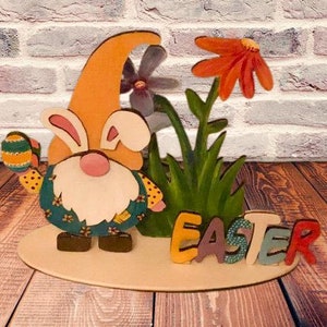 Può includere: Gnomo pasquale di legno che tiene un uovo con un cappello con orecchie di coniglio, in piedi davanti a un cespuglio verde e un fiore con la parola "Pasqua" scritta sotto.
