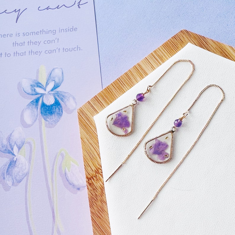 May include: A pair of delicate gold earrings with long chains. Each earring features a teardrop-shaped pendant with a pressed purple flower encased in clear resin. The pendants are accented with a small purple gemstone.