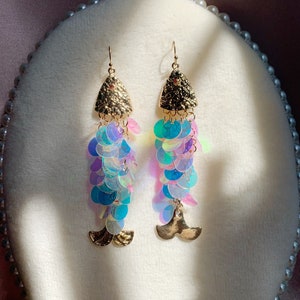May include: A pair of mermaid-themed earrings. The earrings feature gold-colored triangle tops with a small red gem, iridescent sequins in shades of blue, purple, and green, and gold-colored tail charms. The earrings are displayed on a white, oval-shaped surface.