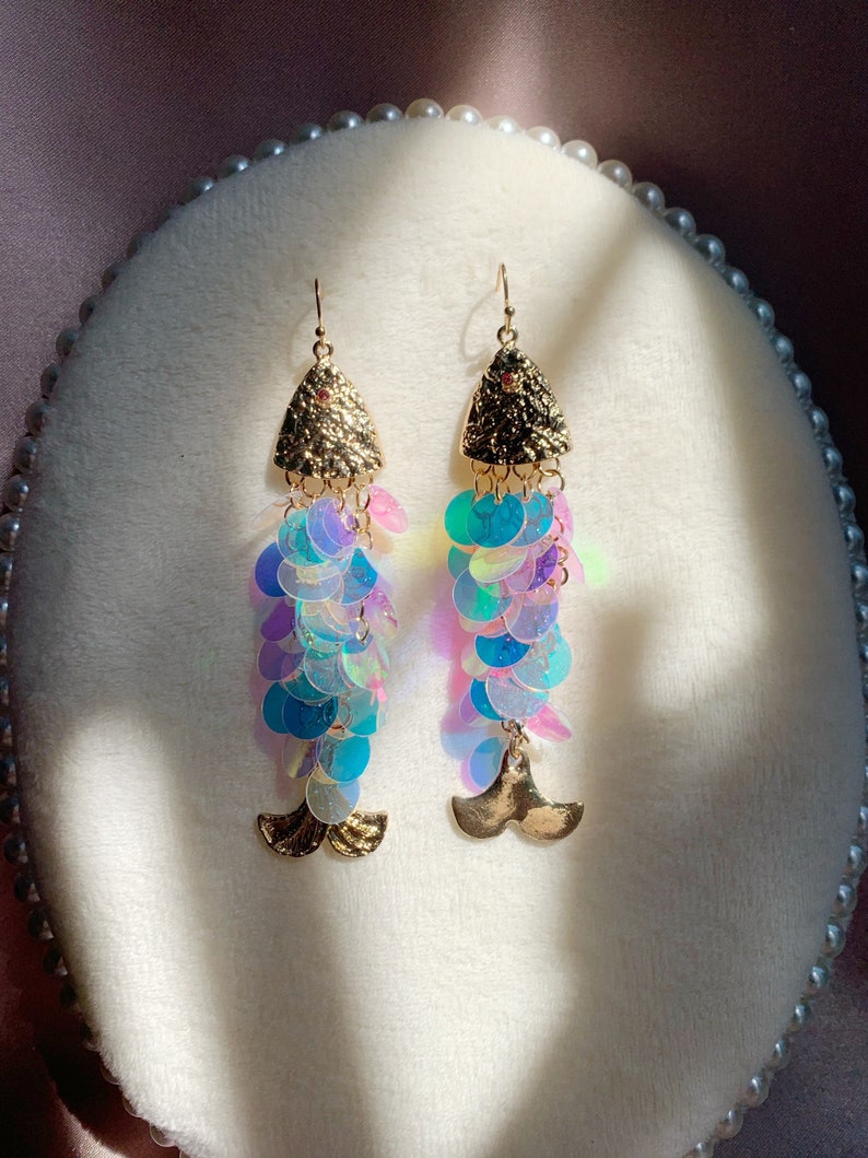 May include: A pair of mermaid-themed earrings. The earrings feature gold-colored triangle tops with a small red gem, iridescent sequins in shades of blue, purple, and green, and gold-colored tail charms. The earrings are displayed on a white, oval-shaped surface.