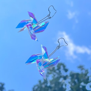 May include: A pair of iridescent pinwheel earrings with silver hooks. The pinwheels are made of a clear plastic material with a holographic finish.