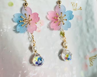 May include: A pair of floral earrings with a delicate design. Each earring features a pink and blue flower with gold accents and a dangling iridescent crystal teardrop. The earrings have a gold hook.