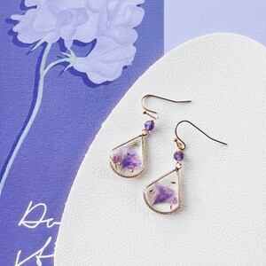 May include: A pair of gold earrings with a teardrop-shaped design. Each earring features a small purple flower pressed inside clear resin and a small purple gemstone bead on the hook.