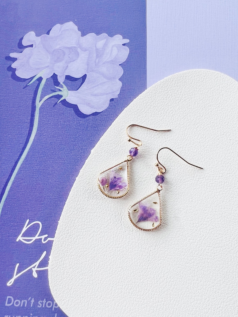 May include: A pair of gold earrings with a teardrop-shaped design. Each earring features a small purple flower pressed inside clear resin and a small purple gemstone bead on the hook.