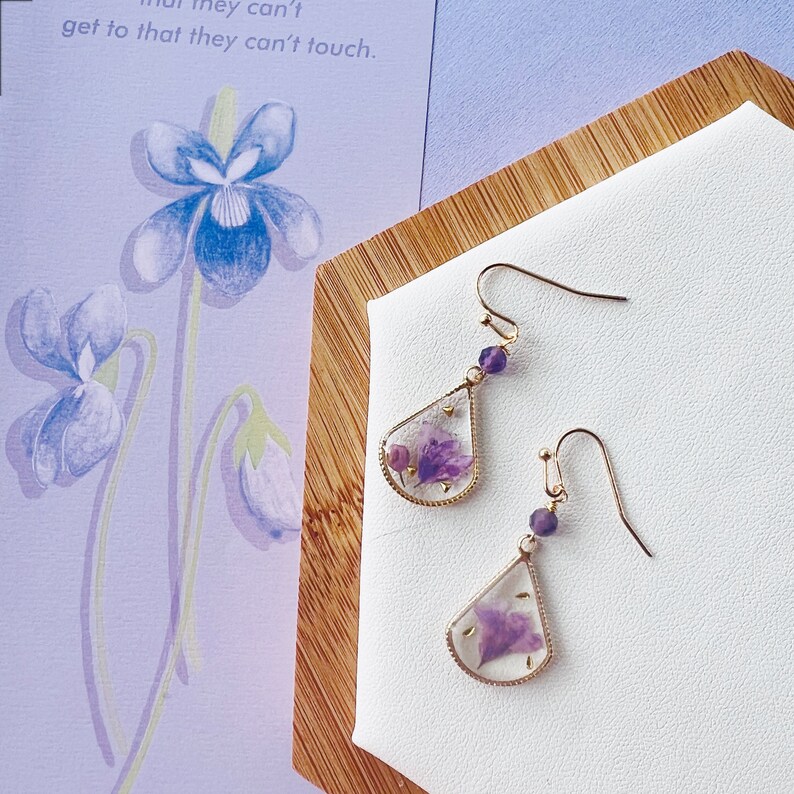 May include: A pair of gold teardrop earrings with purple pressed flowers inside. The earrings are on a white surface with a wooden background.