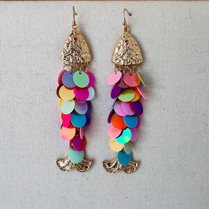 May include: A pair of gold-toned dangle earrings with a fish scale design. The earrings feature a textured triangular top with a small red gem and a cascade of colorful, iridescent sequins in shades of pink, purple, blue, green, and orange.