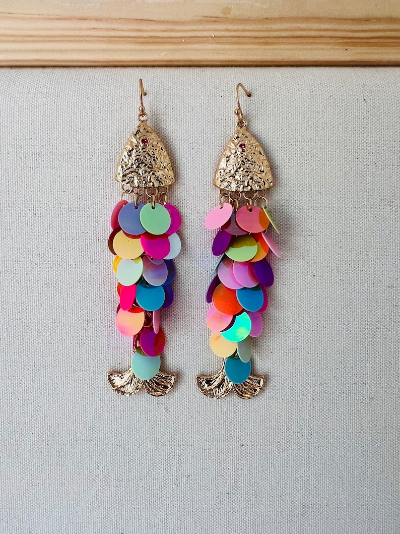 May include: A pair of gold-toned dangle earrings with a fish scale design. The earrings feature a textured triangular top with a small red gem and a cascade of colorful, iridescent sequins in shades of pink, purple, blue, green, and orange.