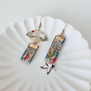 May include: A pair of gold earrings with a blue and white fan design and a white crane charm. The fan design has a red circle in the center. The earrings are hanging from a white surface.