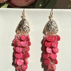 May include: A pair of dangle earrings with a gold-colored top and a cascade of pink, round, shell-like beads. The earrings have a gold-colored fish tail design at the bottom. The background includes pink flowers.