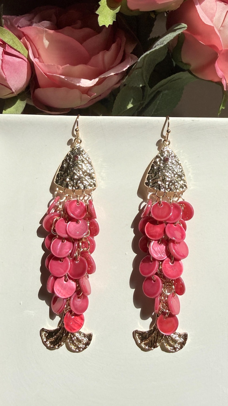 May include: A pair of dangle earrings with a gold-colored top and a cascade of pink, round, shell-like beads. The earrings have a gold-colored fish tail design at the bottom. The background includes pink flowers.
