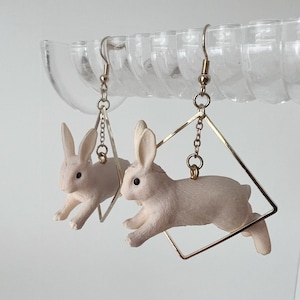 May include: A pair of gold hoop earrings with a white rabbit charm dangling from each hoop. The rabbits are in a jumping pose.