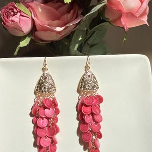 May include: A pair of dangle earrings with a gold-colored top section encrusted with small, shiny stones. The earrings have a cascade of pink, circular beads, and a gold-colored, leaf-like bottom.