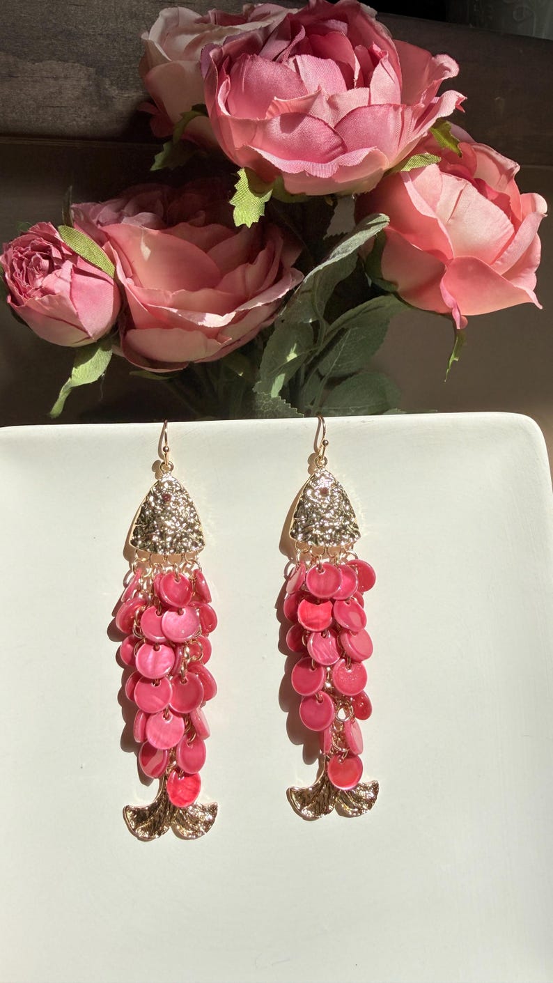 May include: A pair of dangle earrings with a gold-colored top section encrusted with small, shiny stones. The earrings have a cascade of pink, circular beads, and a gold-colored, leaf-like bottom.
