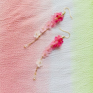Puede incluir: Un par de pendientes largos y delicados con un degradado de flores rosas y blancas que cuelgan de cadenas doradas.