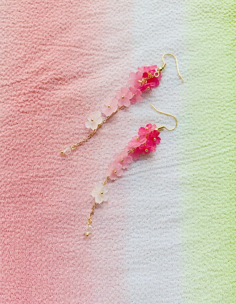 Puede incluir: Un par de pendientes largos y delicados con un degradado de flores rosas y blancas que cuelgan de cadenas doradas.