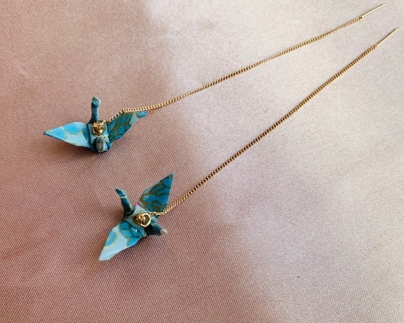 Może przedstawiać: Para długich, złotych kolczyk&oacute;w z łańcuszkiem, z wiszącymi niebieskimi i białymi żurawiami origami.