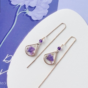 May include: A pair of long, delicate gold chain earrings with a teardrop-shaped pendant featuring a pressed purple flower encased in clear resin. Each pendant has a small purple bead at the top.