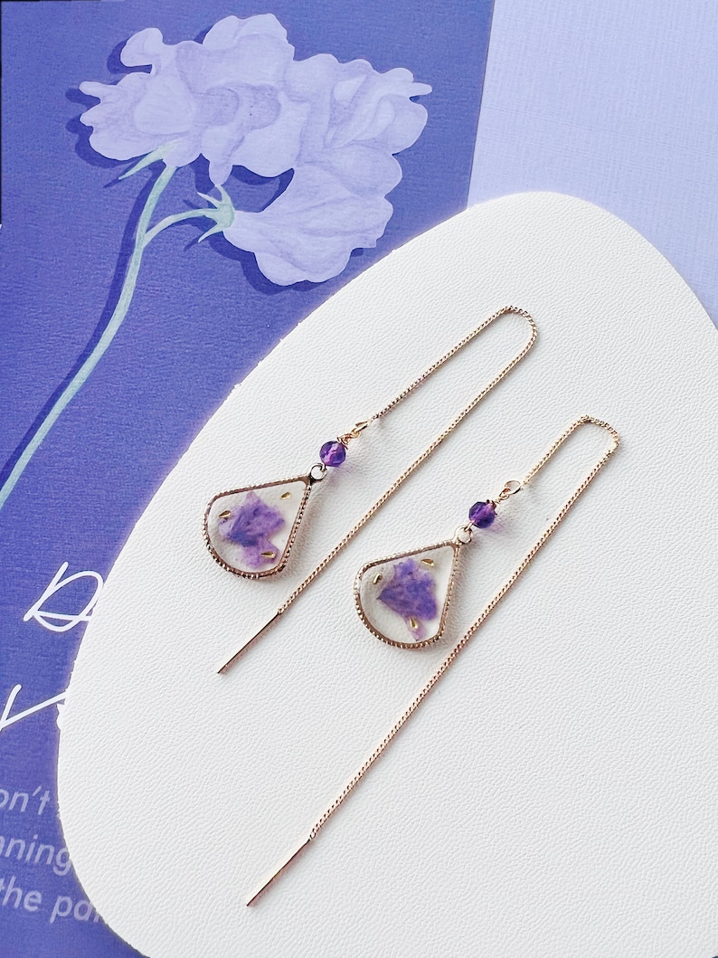 May include: A pair of long, delicate gold chain earrings with a teardrop-shaped pendant featuring a pressed purple flower encased in clear resin. Each pendant has a small purple bead at the top.