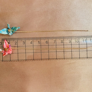 Może przedstawiać: Dwa złote łańcuszki z przymocowanymi do nich origami z żurawiami. Łańcuszki są mierzone za pomocą linijki z oznaczeniami od 0 do 13 cali.