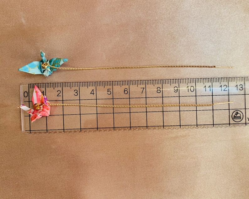 Może przedstawiać: Dwa złote łańcuszki z przymocowanymi do nich origami z żurawiami. Łańcuszki są mierzone za pomocą linijki z oznaczeniami od 0 do 13 cali.
