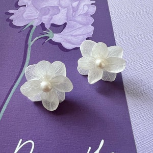 May include: Two white flower-shaped earrings with a pearl center rest on a purple card with a floral illustration and the word "Don't" in white script. The earrings have delicate, translucent petals.