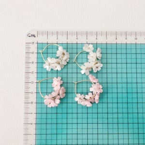 May include: Four pairs of hoop earrings. Two pairs feature white flower clusters, and two pairs have pink flower clusters. The hoops are gold-colored. The earrings are displayed on a grid with centimeter markings.
