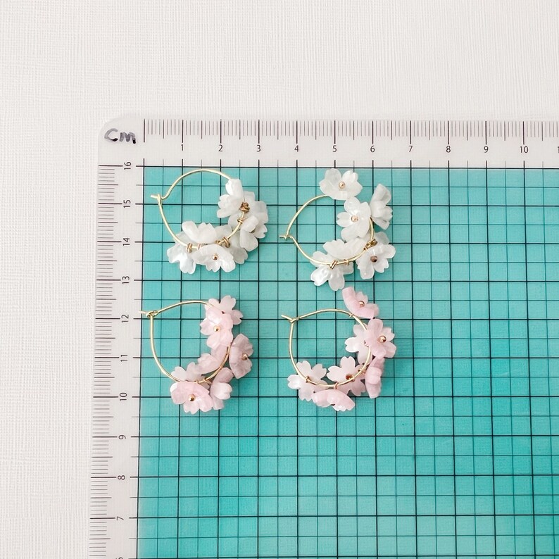 May include: Four pairs of hoop earrings. Two pairs feature white flower clusters, and two pairs have pink flower clusters. The hoops are gold-colored. The earrings are displayed on a grid with centimeter markings.