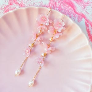 Op de afbeelding: Delicate roze bloemen oorbellen met goudkleurige kettingen en parelmoer kralen. De oorbellen hebben doorschijnende roze bloemblaadjes en worden gepresenteerd op een roze bord.